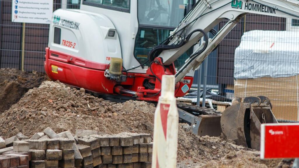 An excavator working at a construction site in a suburban neighborhood, framed by safety barriers.